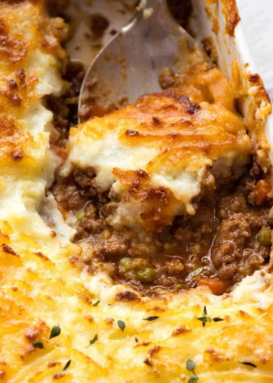 A spoonful of shepherd's pie lifted from the dish, showing lamb and pea filling under golden, cheesy mash