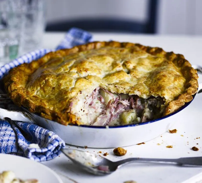 A golden double-crust pie in an enamel dish, cut open to show shredded ham, potato and a creamy sauce