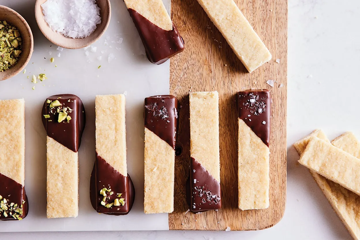 Shortbread strips on a board, some half-dipped in dark chocolate and topped with flaky salt or chopped pistachios