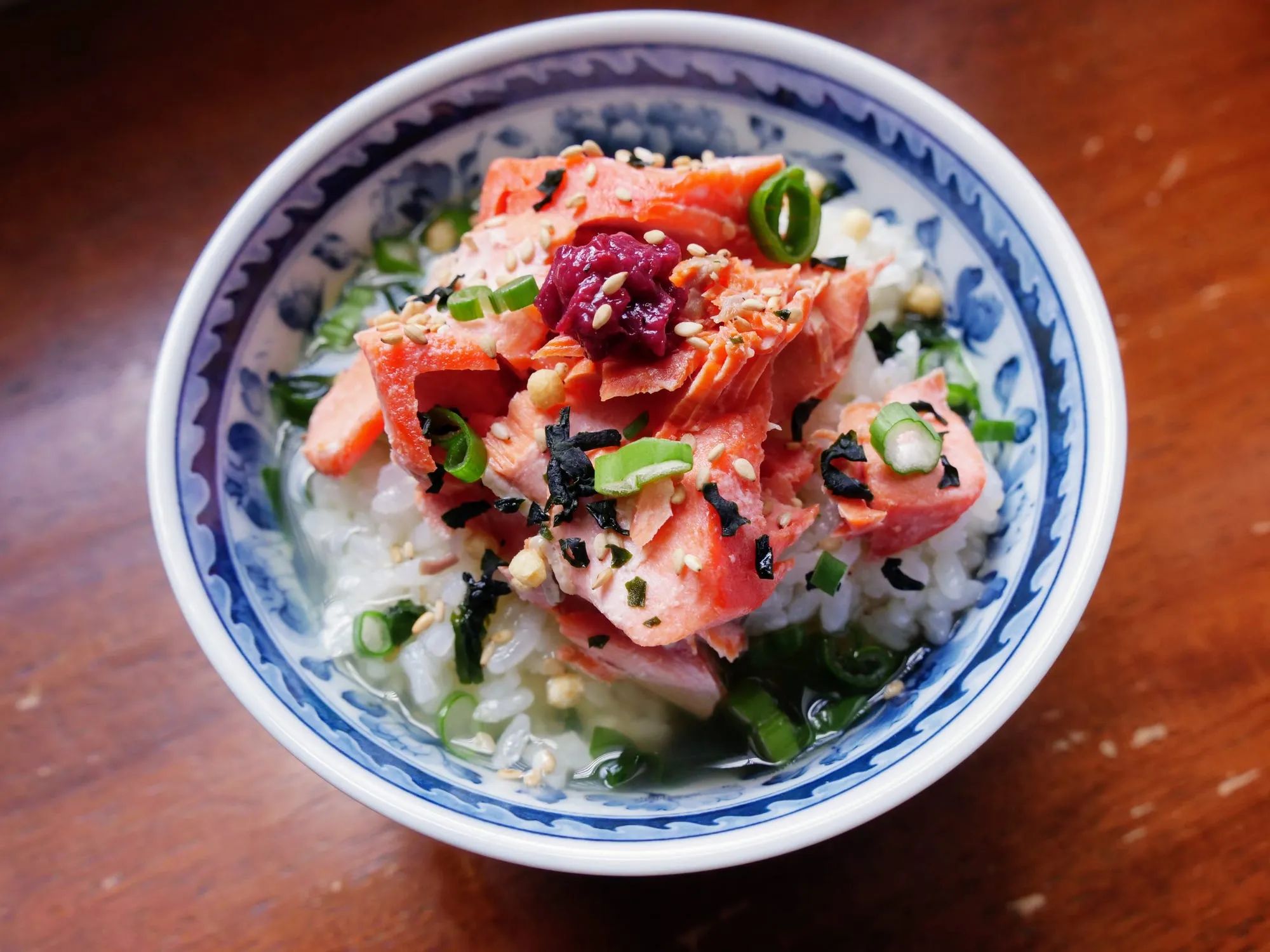 A blue and white bowl of rice in green tea, topped with flaked salmon, umeboshi, sesame seeds, nori and spring onion