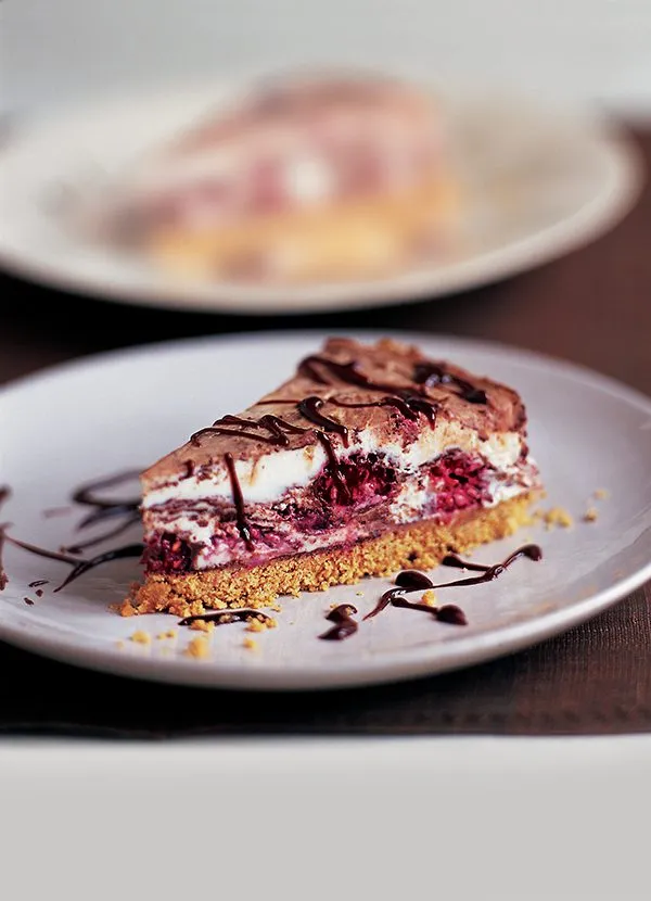 A slice of cheesecake with a biscuit base, raspberries and chocolate swirls, drizzled with melted chocolate